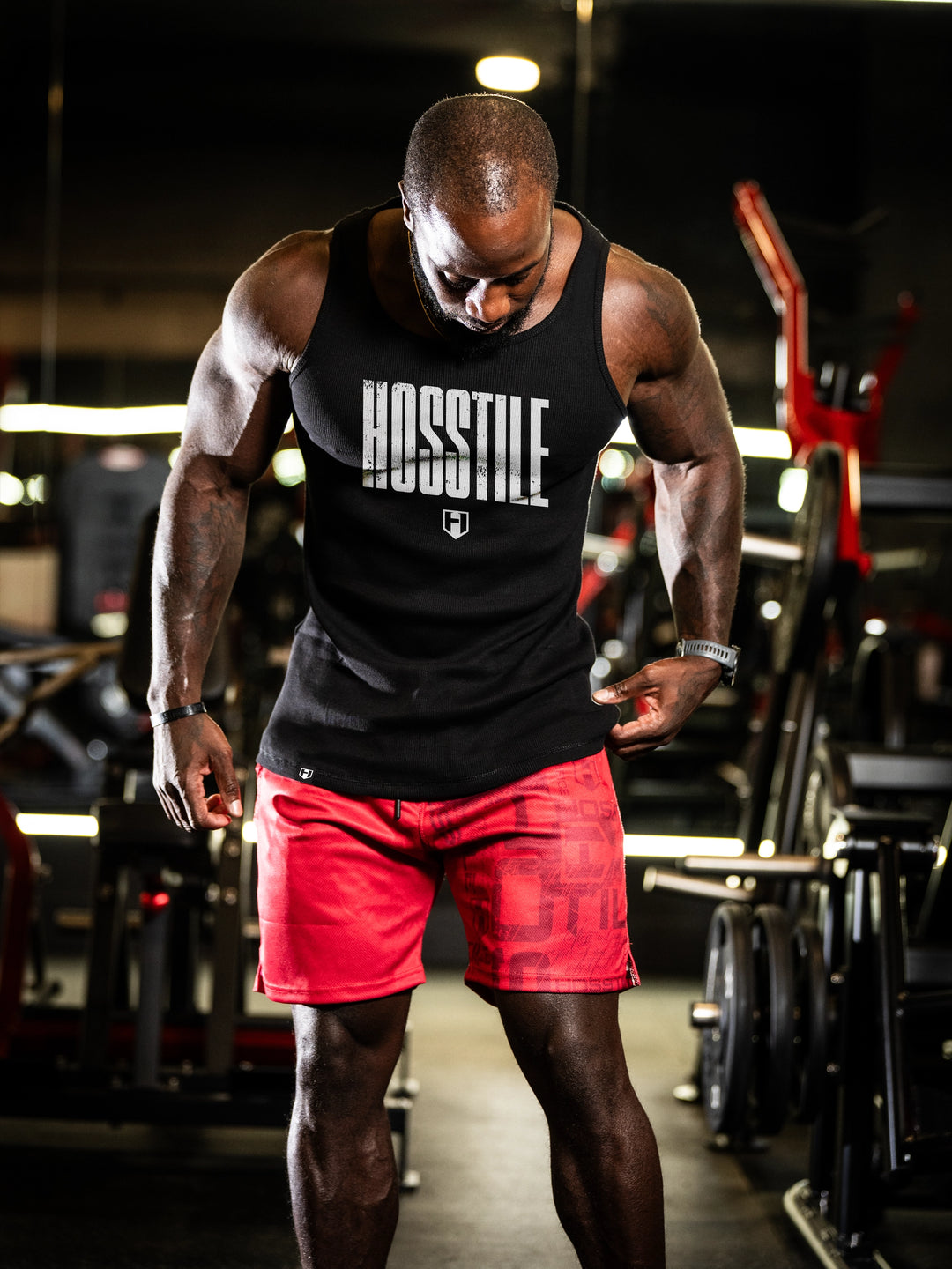 Man wearing a black tank top with 'HOSSILE' text and red shorts in a gym setting#color_black