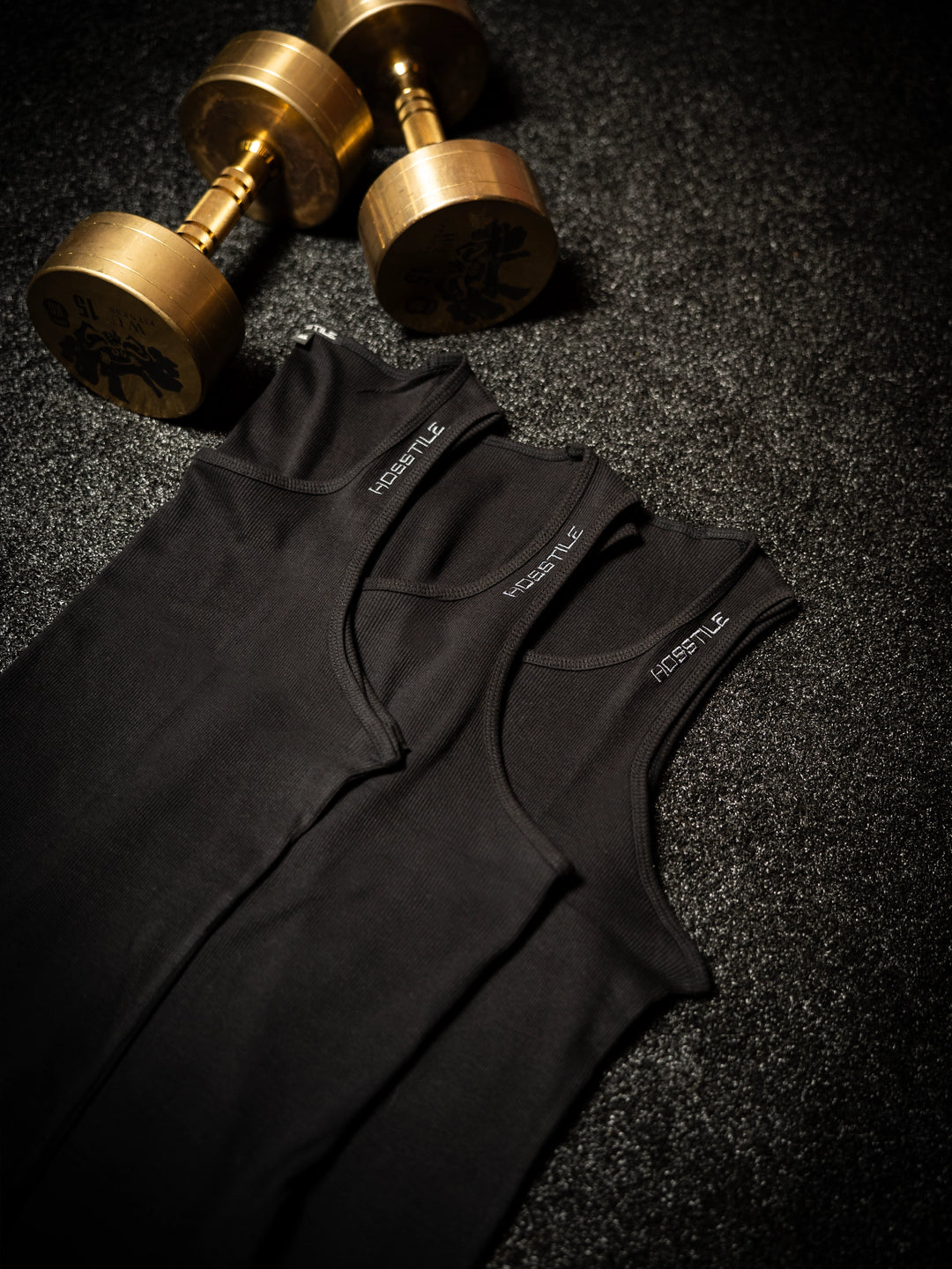 Black sleeveless shirt with Hosstile branding on a dark textured surface with gold dumbbells#color_black