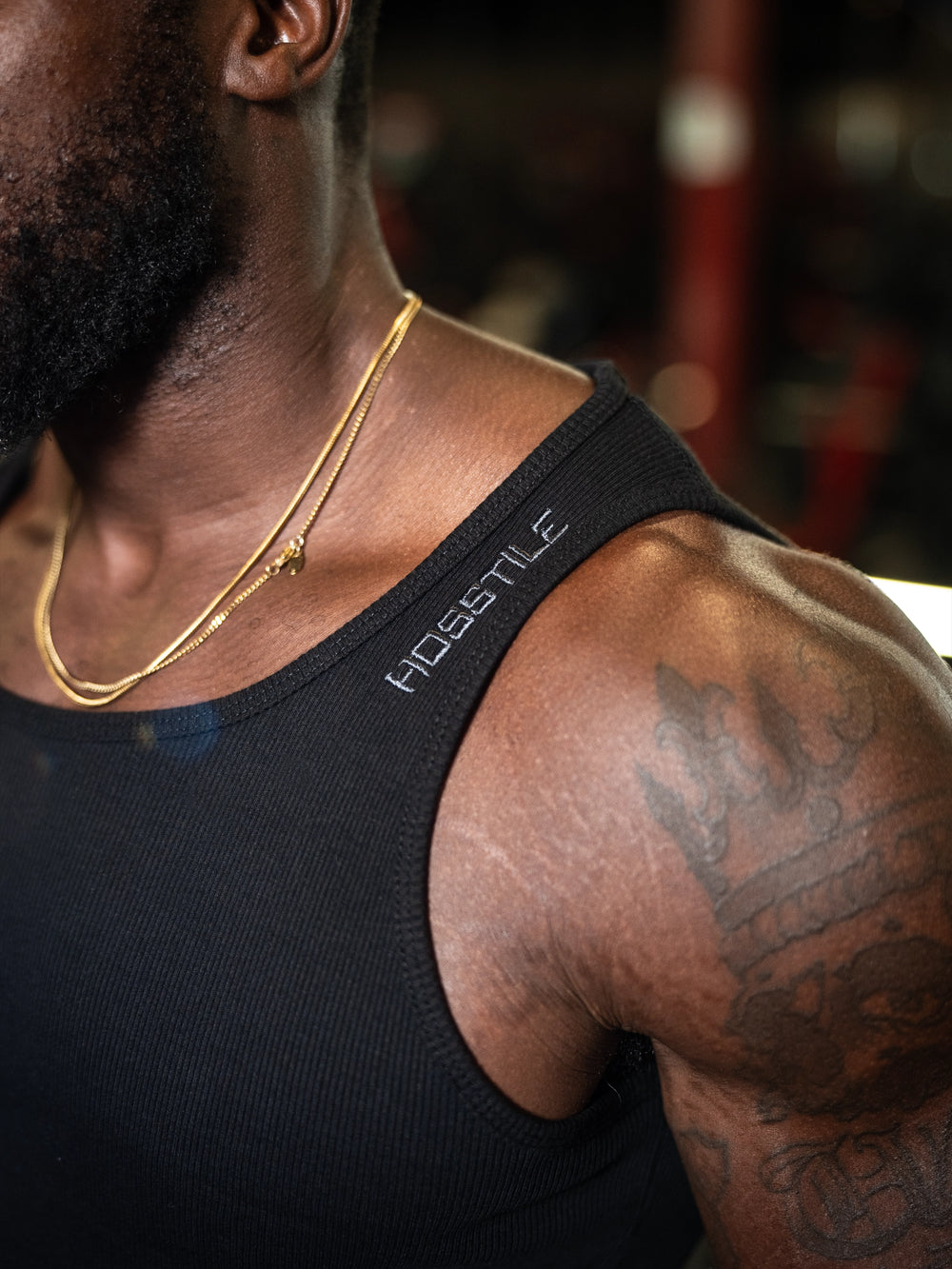 Person wearing a black tank top with Hosstile branding, gold necklace, and tattoos#color_black