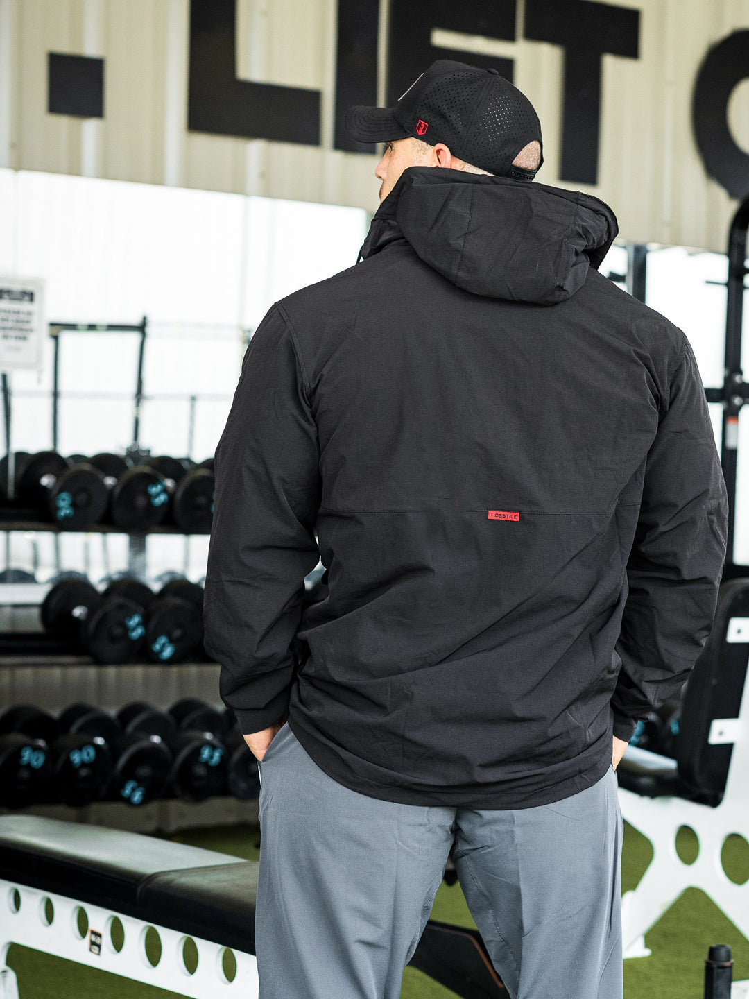 Bodybuilder Martin Fitzwater in a gym wearing a black hoodie windbreaker jacket and dark pants with visible branding#color_black