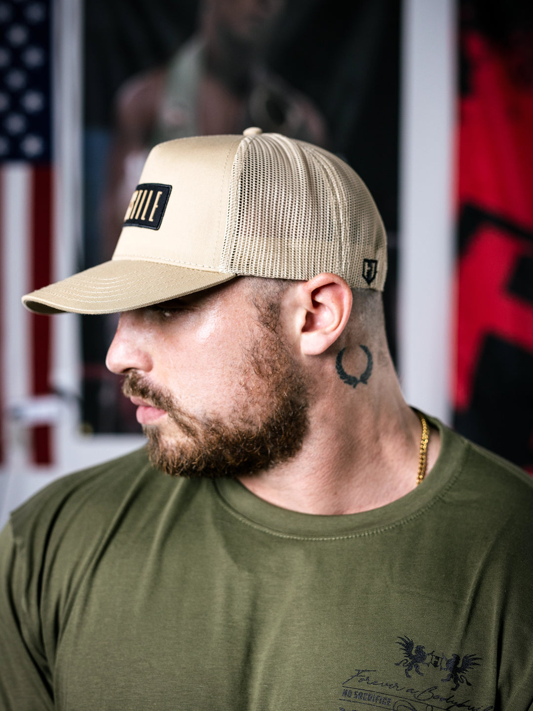 IFBB Martin Fitzwater wearing a sand trucker hat with 'Hosstile' logo and a green t-shirt with graphic design.#color_desert-sand