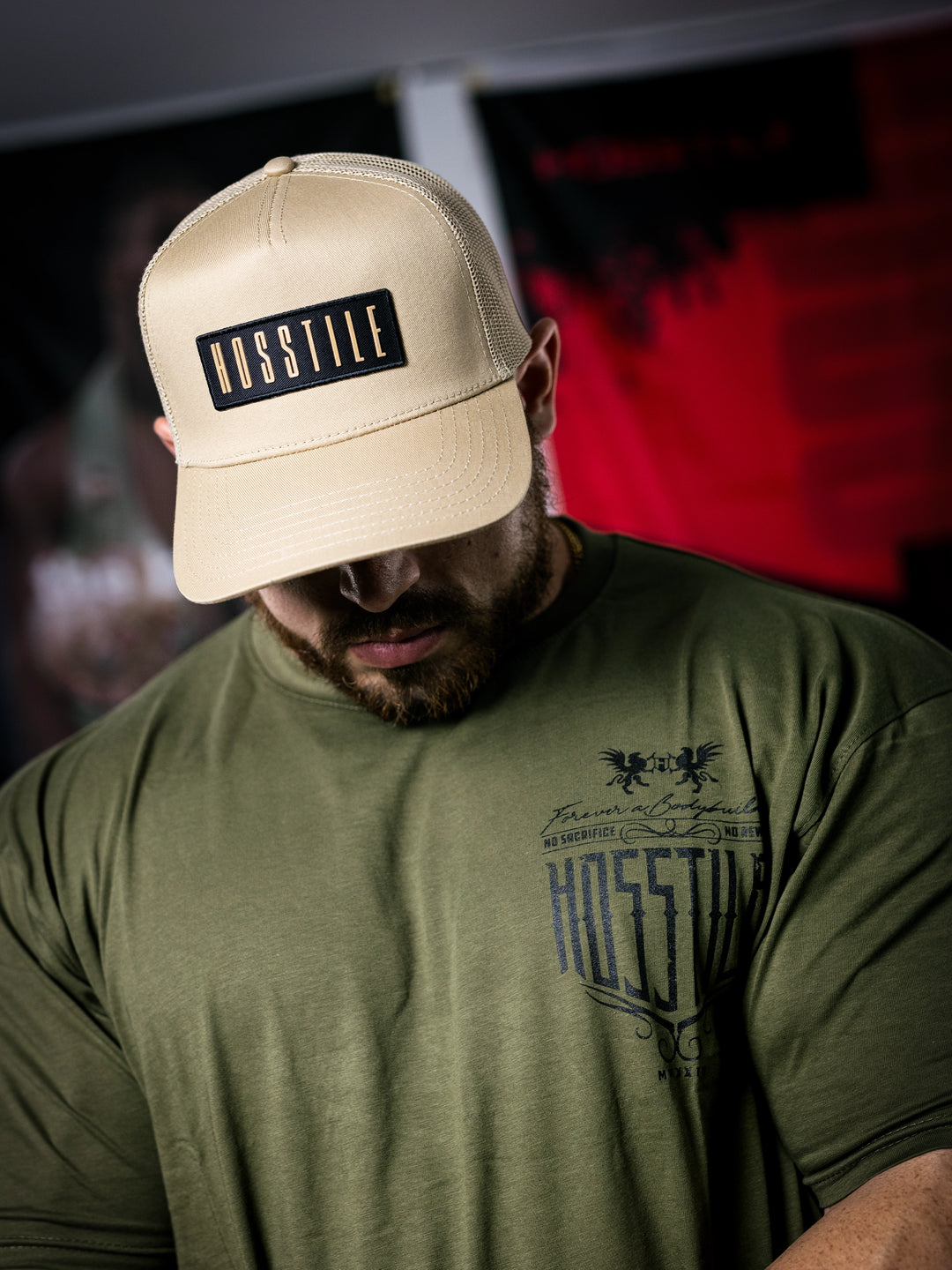 IFBB Martin Fitzwater wearing a sand trucker hat with 'Hosstile' logo and a green t-shirt with graphic design.#color_desert-sand