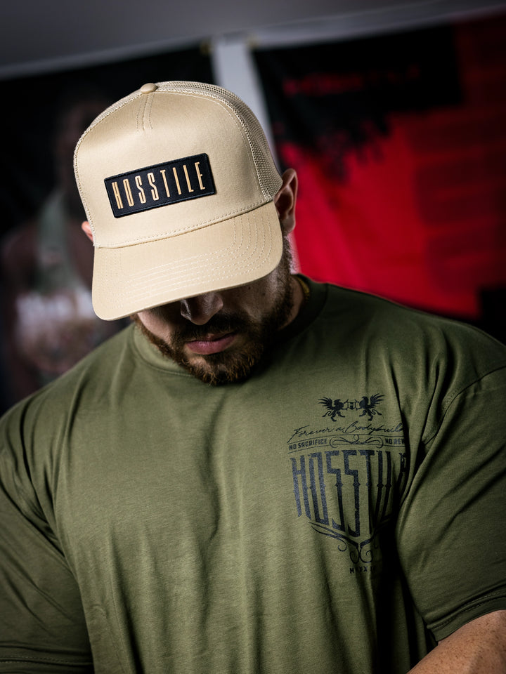 IFBB Martin Fitzwater wearing a sand trucker hat with 'Hosstile' logo and a green t-shirt with graphic design.#color_desert-sand