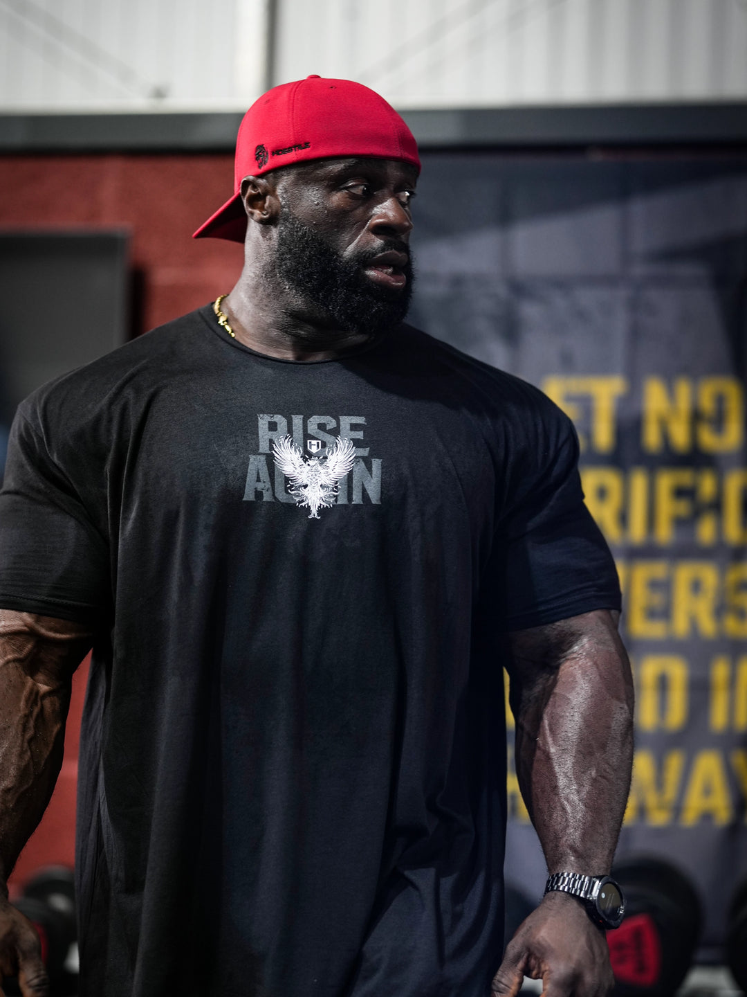 Samson Dauda wearing a black t-shirt with a logo and text, red cap, and watch, standing outdoors#color_black