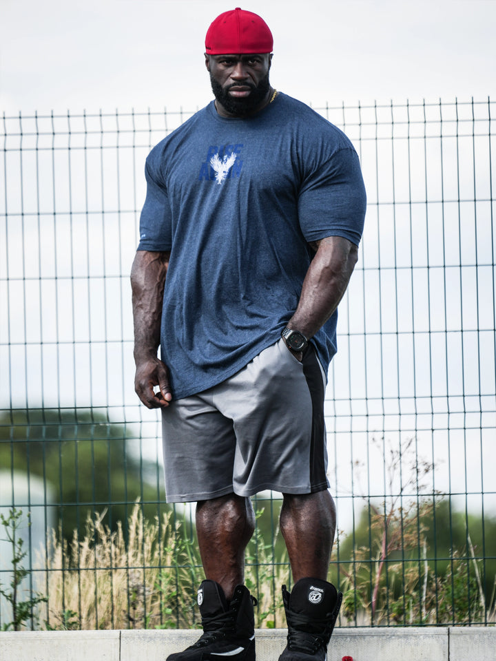 Samson Dauda wearing a blue t-shirt with a logo, gray shorts, and black shoes, standing outdoors with a wire fence and greenery in the background#color_navy