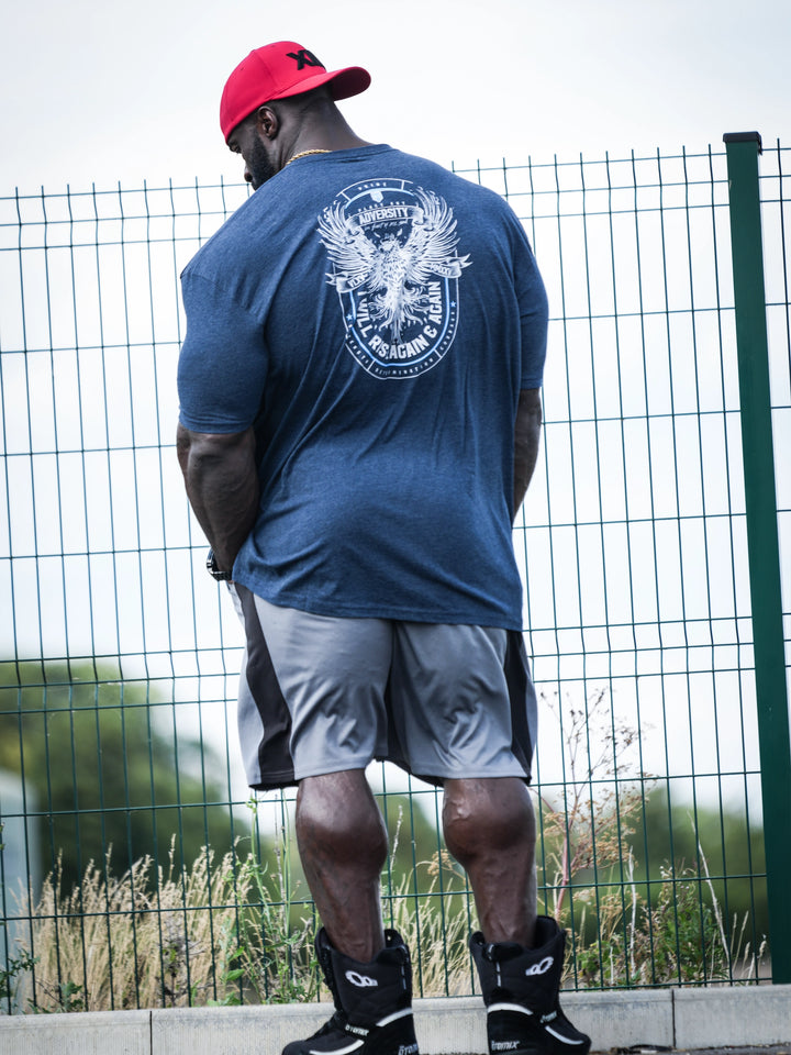 Samson Dauda wearing a blue t-shirt with a logo, gray shorts, and black shoes, standing in front of a wire fence#color_navy