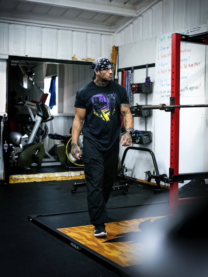 Bodybuilder Ben Chow wearing a black t-shirt with a graphic design and a cap, standing in a gym.#color_black-african-sunrise