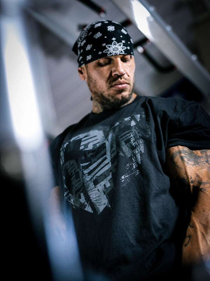 Bodybuilder Ben Chow wearing a black t-shirt with a graphic design and a cap, standing in a gym.#color_black-grey
