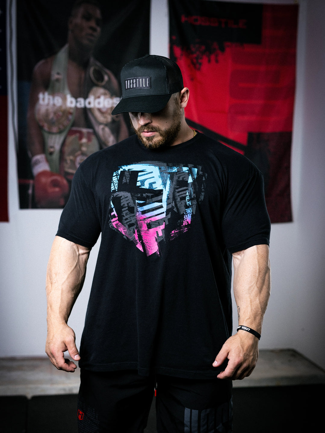 IFBB Pro Bodybuilder Martin Fitzwater wearing a black t-shirt with a graphic design and a cap, standing in a room with boxing-themed decor.#color_black-miami-vice