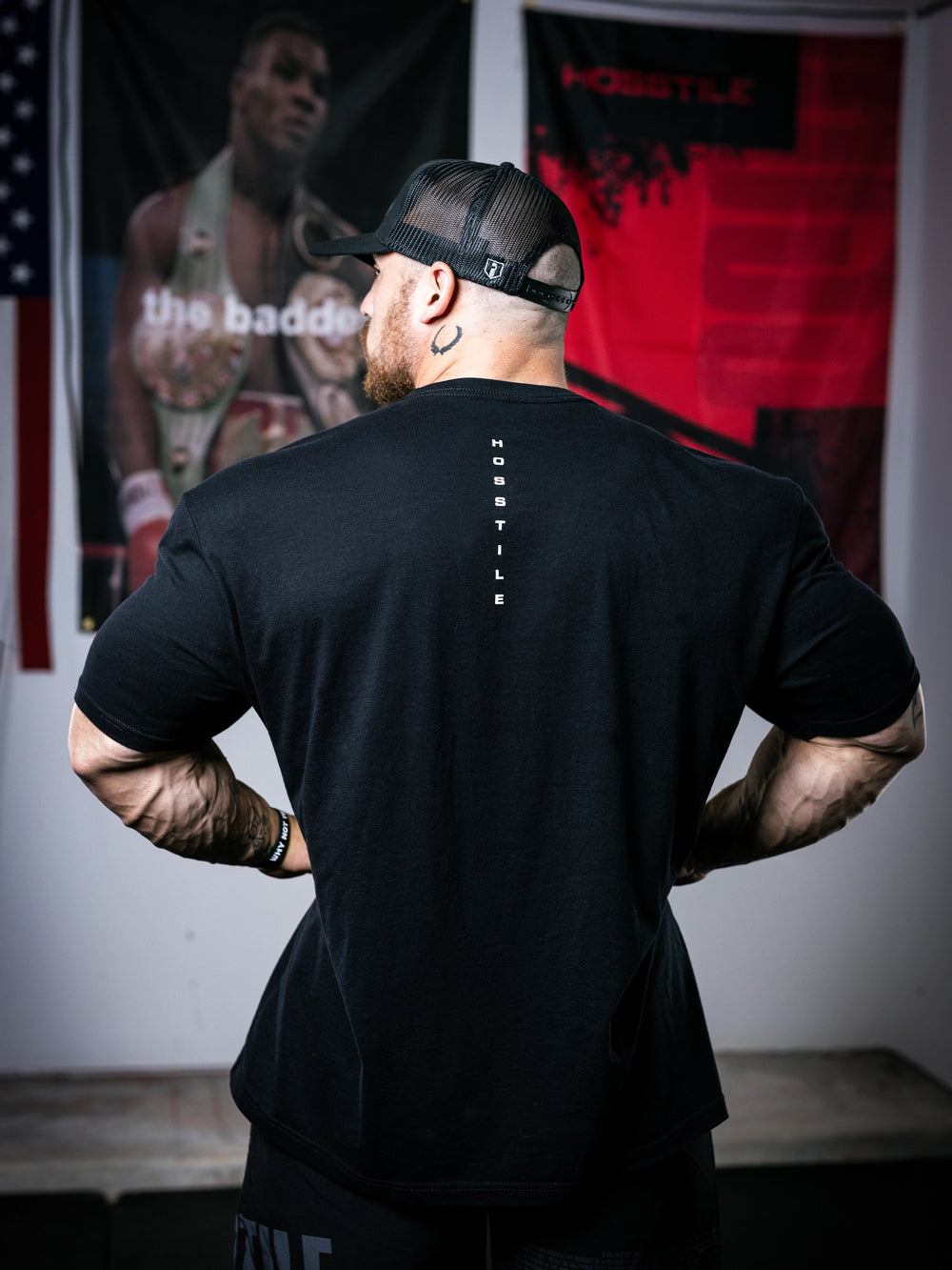 IFBB Pro Bodybuilder Martin Fitzwater wearing a black t-shirt with a graphic design and a cap, standing in a room with boxing-themed decor.#color_black-miami-vice