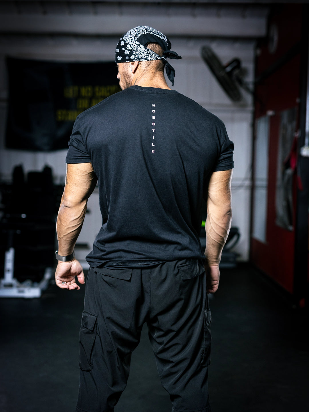 Bodybuilder Ben Chow wearing a black t-shirt with a graphic design and a cap, standing in a gym.#color_black-ocean