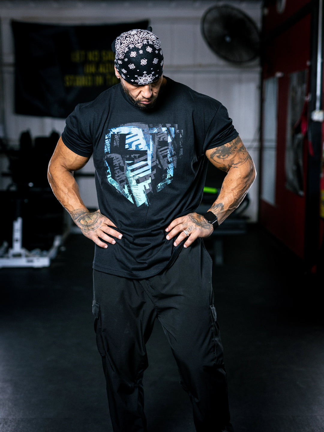Bodybuilder Ben Chow wearing a black t-shirt with a graphic design and a cap, standing in a gym.#color_black-ocean