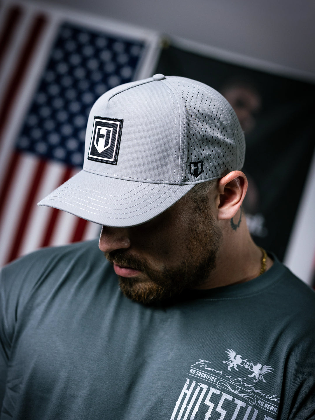 Man wearing a white cap with a logo, looking down, with an American flag in the background.#color_grey
