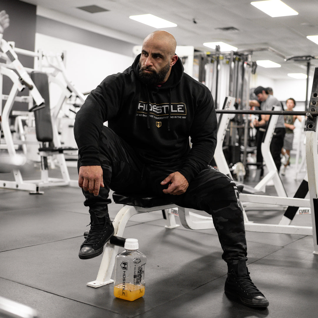 Bodybuilder Fouad Abiad wearing Hosstile Signature Hoodie sitting on a bench in the gym#color_black